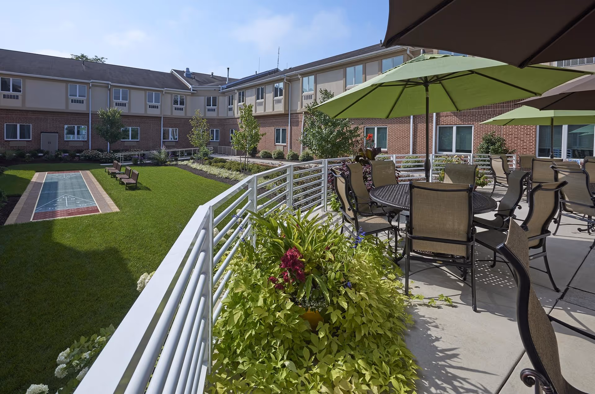 Outdoor patio area at Lutheran Community at Telford with tables, chairs, and large green umbrellas. The patio overlooks a grassy courtyard with benches, small trees, and a shuffleboard court. The building surrounds the courtyard with multiple windows visible.