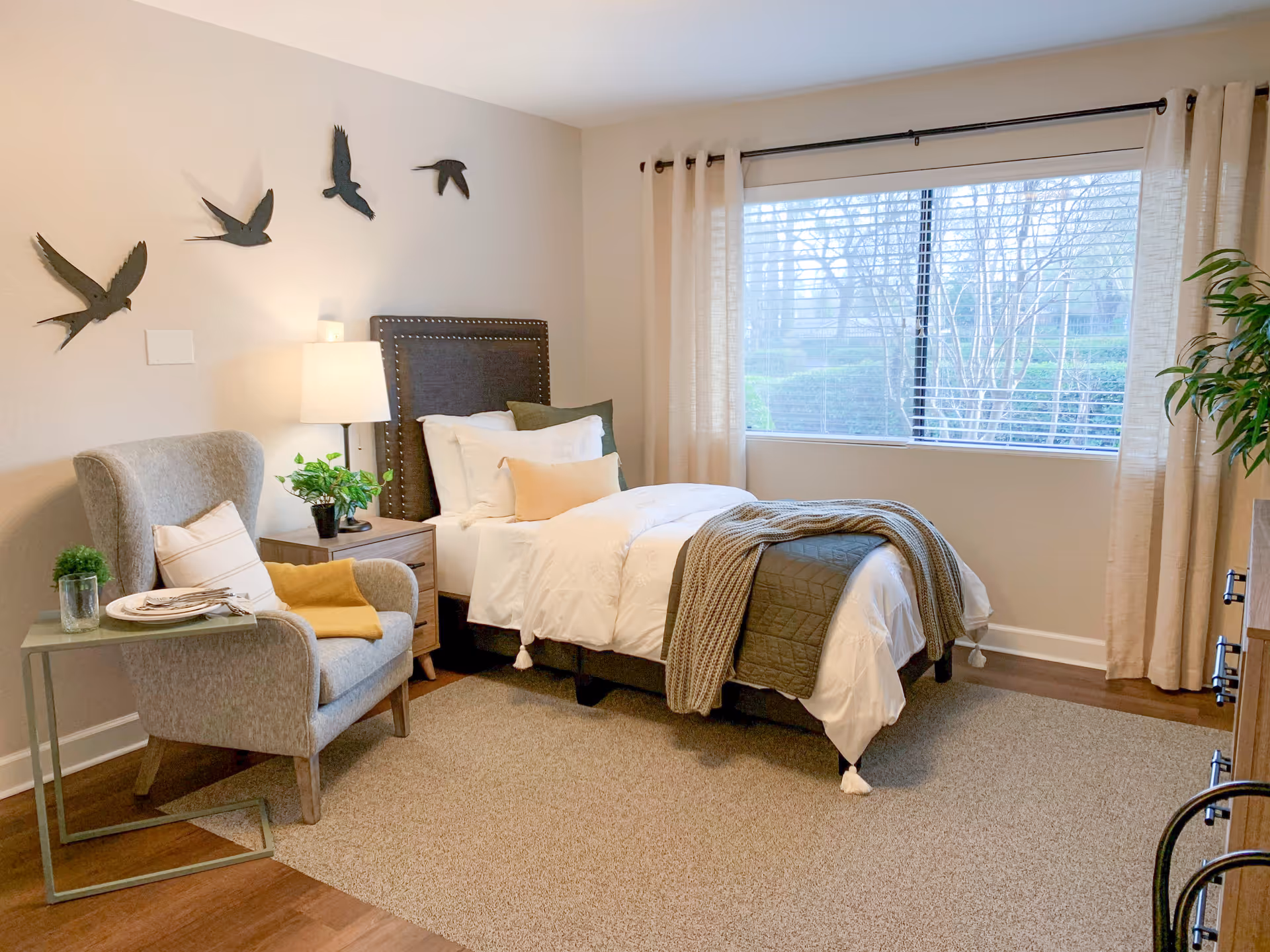 A cozy bedroom with a single bed dressed in white and green bedding, a gray upholstered headboard, and a knitted throw blanket. Next to the bed is a wooden nightstand with a lamp and a small plant. A comfortable gray armchair with a pillow and a yellow throw is positioned beside a small side table holding a glass and a plate. The room features a large window with beige curtains and a view of trees outside. Decorative bird wall art is mounted above the nightstand.