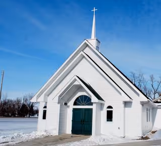 White church-like building with green double doors and a tall steeple topped by a cross, surrounded by snow under a blue sky.
