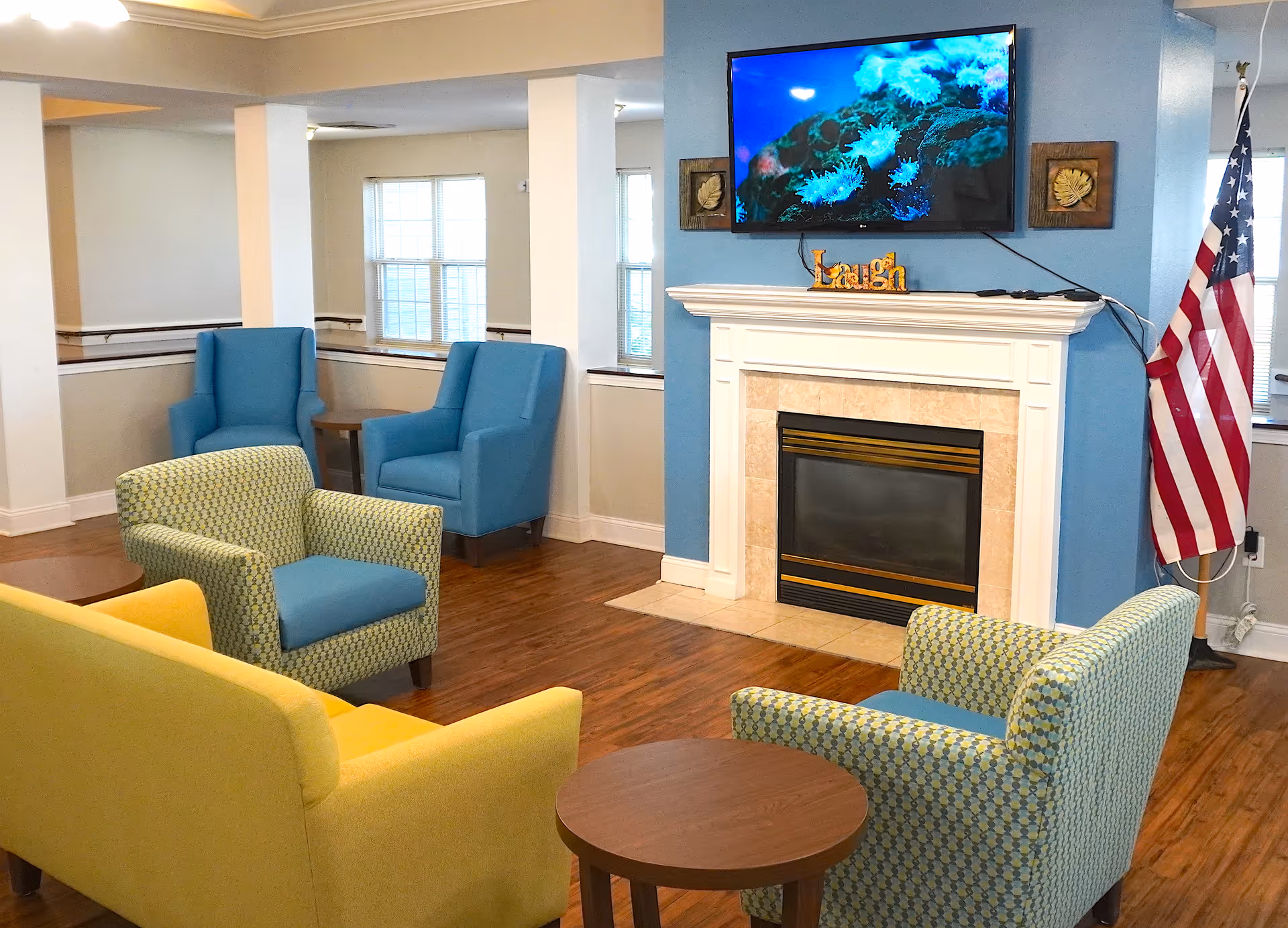 A cozy living room area with a yellow sofa, two patterned armchairs, and two blue armchairs arranged around a wooden coffee table. There is a fireplace with a white mantel and a TV mounted above it displaying an underwater scene. An American flag stands to the right of the fireplace.