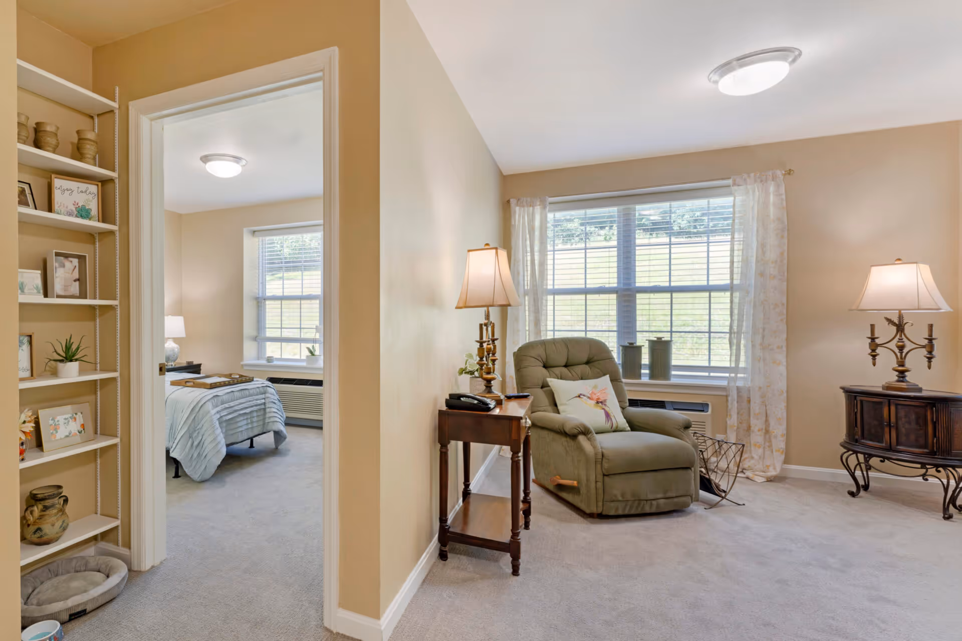 A cozy interior view of a senior living facility showing a living room with a green recliner chair, two table lamps on wooden side tables, and a large window with sheer curtains letting in natural light. To the left, there is an open doorway leading to a bedroom with a bed, nightstand, and window. A shelving unit with decorative items and framed pictures is visible on the left side of the image.