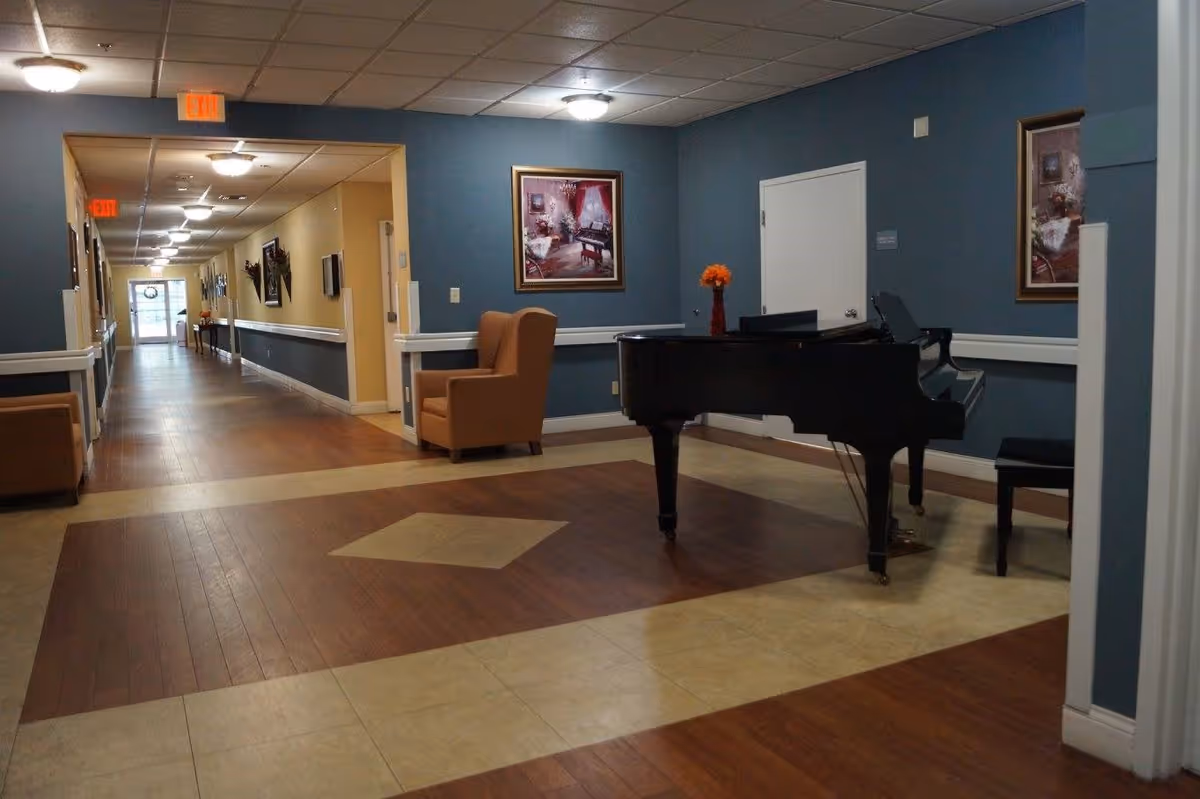 Interior hallway of a senior living facility with wood and tile flooring, a black grand piano with a vase of orange flowers on top, beige armchairs, framed paintings on blue walls, and ceiling lights. The hallway extends into the distance with exit signs visible.