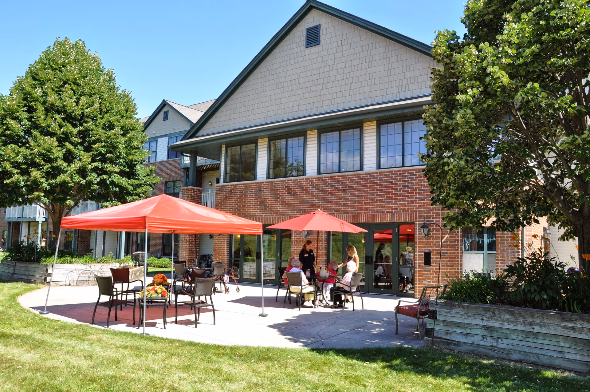 Outdoor patio area at a senior living facility with red umbrellas providing shade over tables and chairs. Several people are seated and standing, engaging in conversation. The building has a brick lower facade with large windows and a gabled roof. There are trees and greenery surrounding the patio.