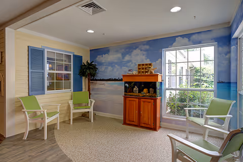 A cozy seating area with four white chairs featuring green cushions arranged around a carpeted circular area. The walls are decorated with a beach-themed mural showing a sandy shore, ocean, and blue sky with clouds. There is a wooden cabinet with a fish tank on top, and a window with blue shutters on one wall. A potted plant is placed near the corner, and natural light comes through a large window overlooking greenery outside.