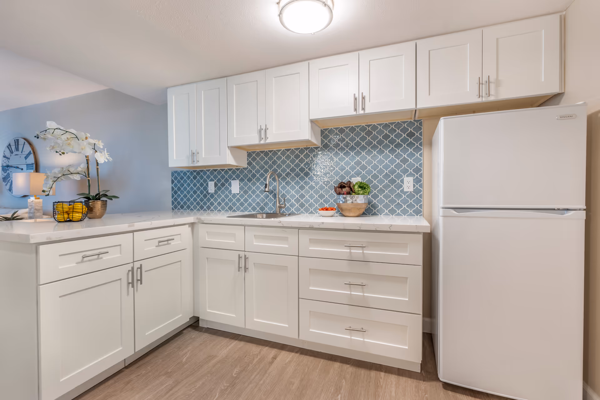 Bright modern kitchenette with white cabinets, a blue patterned tile backsplash, a sink on a marble countertop, and a white refrigerator.