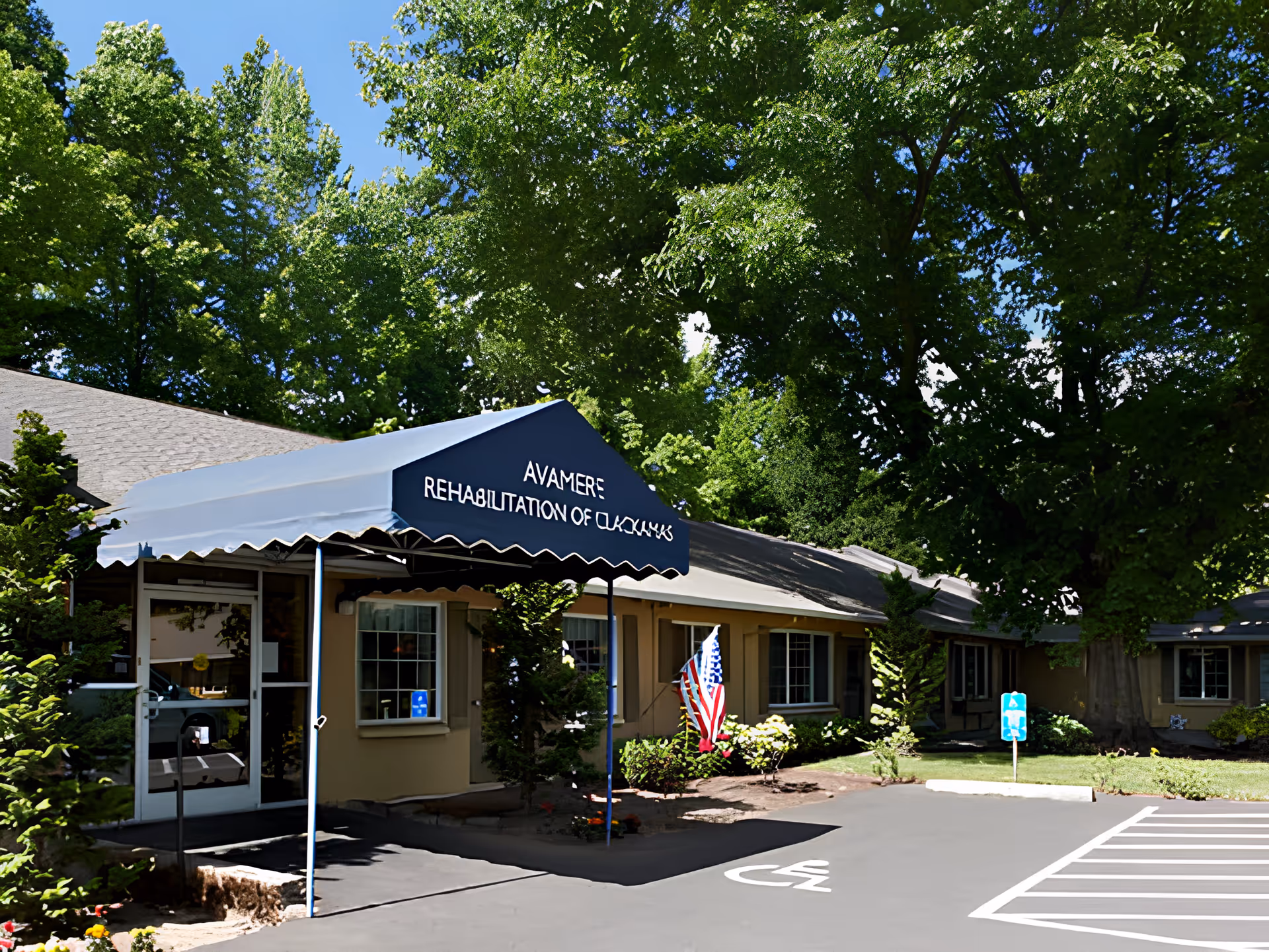 Exterior view of Avamere Rehabilitation of Clackamas building entrance with a blue canopy displaying the facility name. The building is surrounded by green trees and landscaping, with a parking area including a handicapped parking space in front.