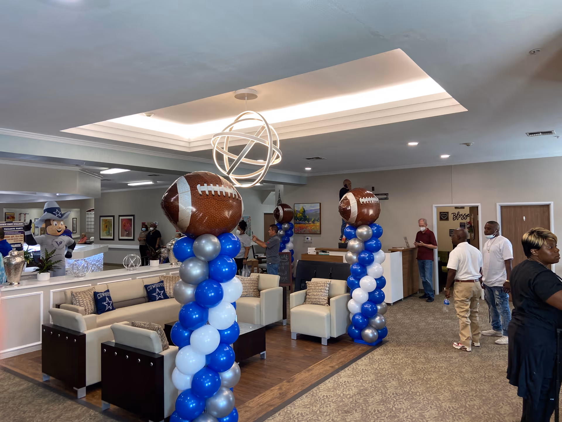 Interior of a senior living facility lobby decorated with blue, white, and silver balloon columns topped with large football-shaped balloons. Several people are standing and walking around the area. There are beige sofas and chairs with Dallas Cowboys pillows, a reception desk, and a large ceiling light fixture. A mascot figure is visible in the background near the reception area.