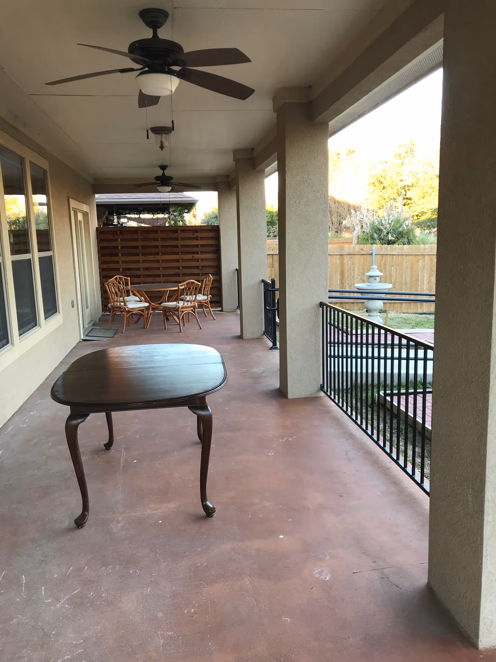 Covered outdoor patio area with a wooden table in the foreground and a set of four wooden chairs with cushions around a small table in the background. The patio has ceiling fans and is enclosed by a railing on one side, with a view of a fenced yard and a decorative fountain outside.