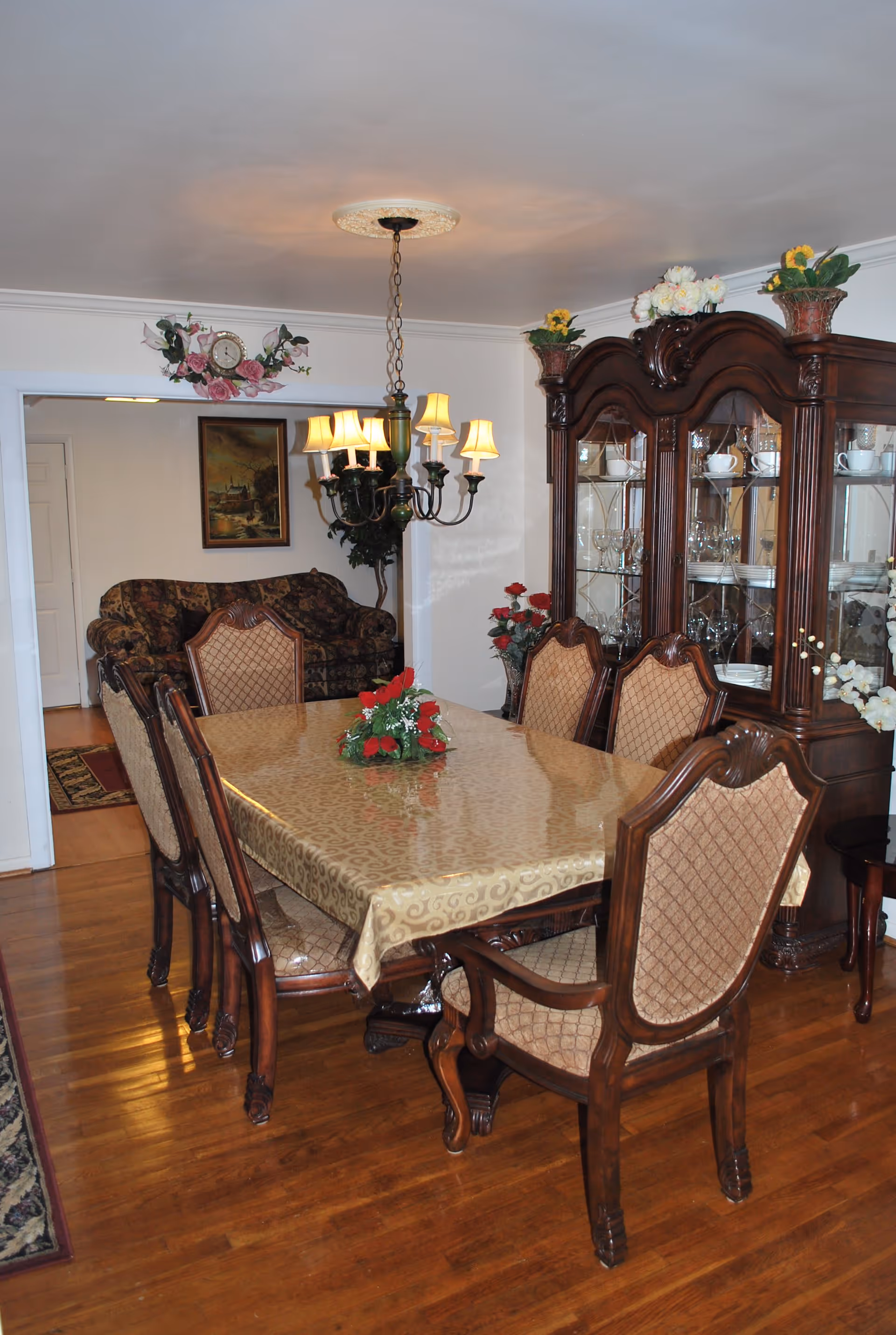 A traditional dining room with a rectangular table covered by a gold patterned tablecloth and surrounded by six upholstered wooden chairs. A chandelier with six lampshades hangs above the table. In the background, there is a wooden china cabinet displaying glassware and dishes, and a floral arrangement on top. A floral clock and a framed painting are mounted on the wall, and a floral-patterned sofa is partially visible through an open doorway.