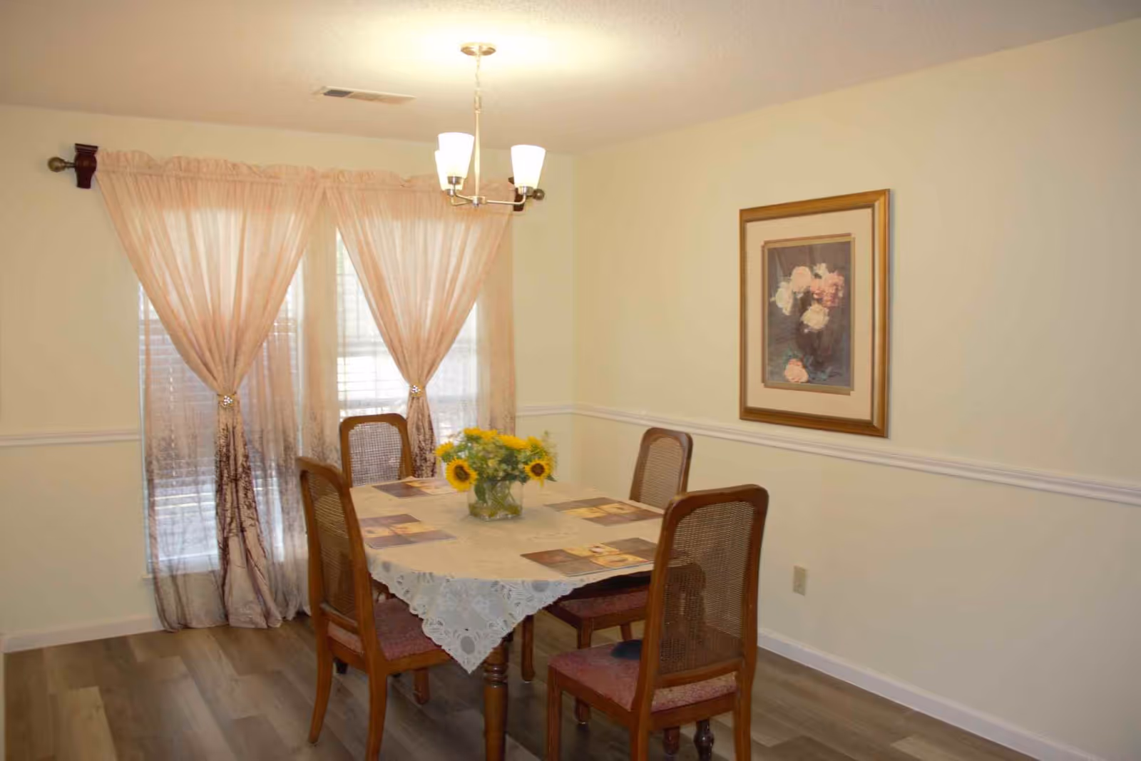 A dining room with a wooden table covered by a lace tablecloth and placemats, surrounded by four wooden chairs with cushioned seats. A vase with sunflowers is placed in the center of the table. Behind the table, there are two windows with sheer pink curtains tied back. A framed floral painting hangs on the wall to the right, and a ceiling light fixture with three lamps is above the table.