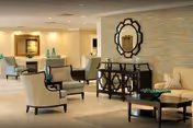 A spacious and well-lit living room area with beige walls and tiled floors. The room features several cushioned armchairs and sofas arranged around dark wooden coffee tables. Decorative elements include a large ornate mirror on the wall, a sideboard with vases, and framed artwork. The overall decor is modern and inviting.