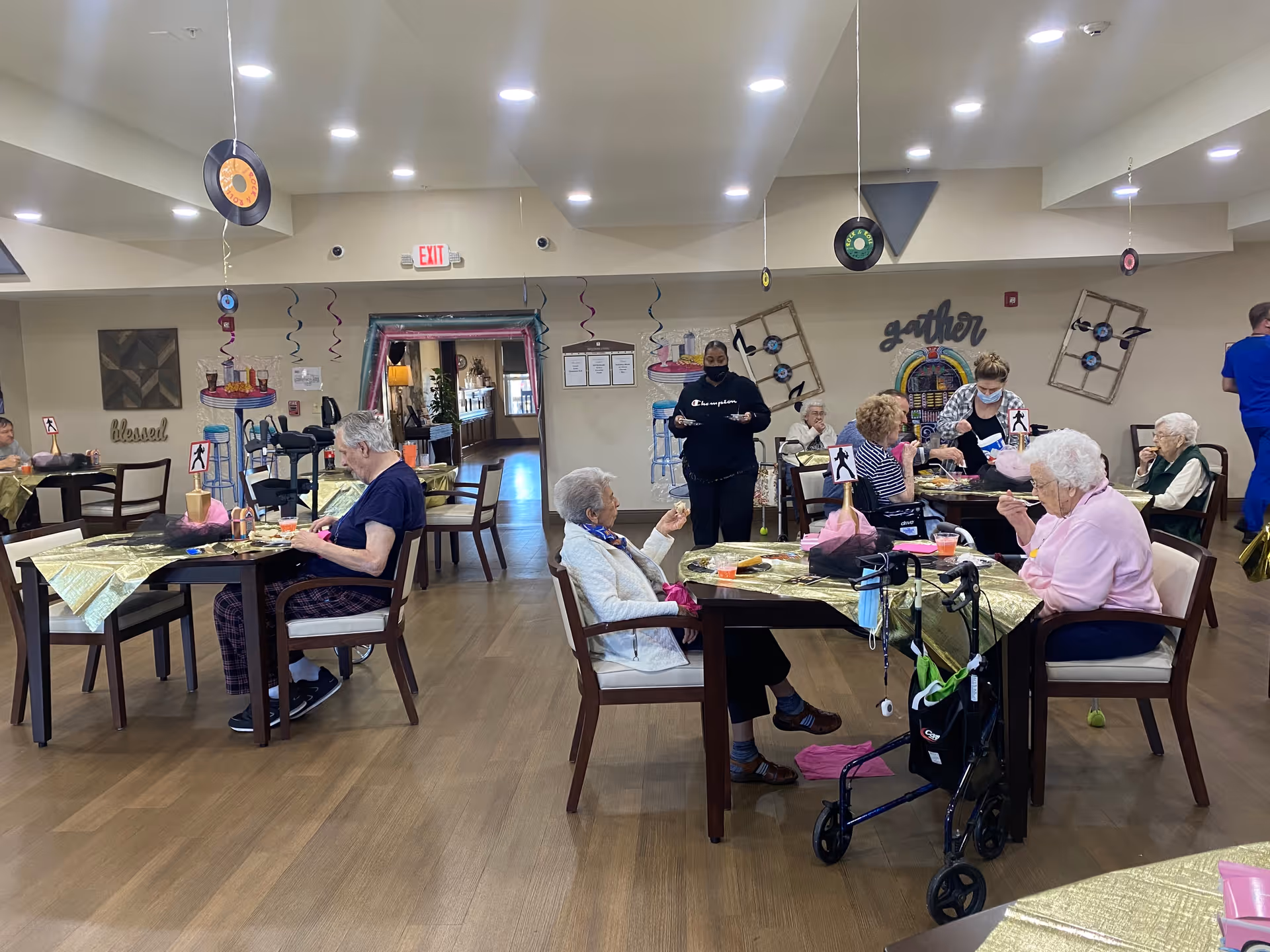Seniors seated at decorated dining tables in a communal dining/activity room with staff assisting.