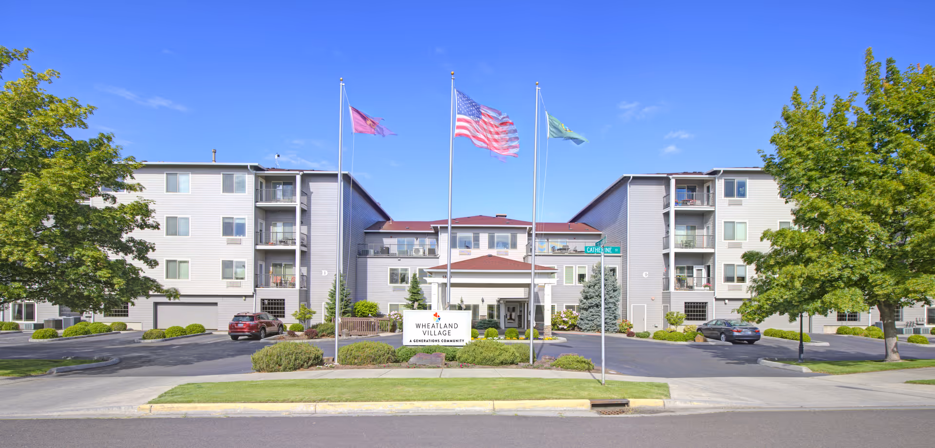 Front exterior view of Wheatland Village a Generations Community building with three flagpoles displaying flags, a parking area with cars, and trees on both sides under a clear blue sky.