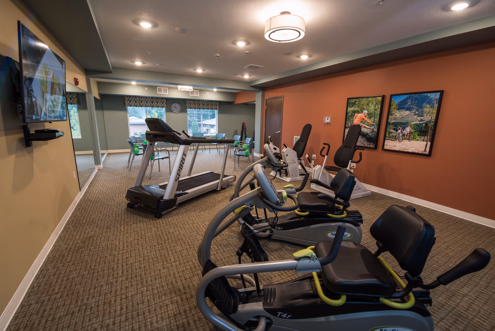 Bright fitness room with treadmills, recumbent exercise bikes, a wall-mounted TV, and a table with chairs.