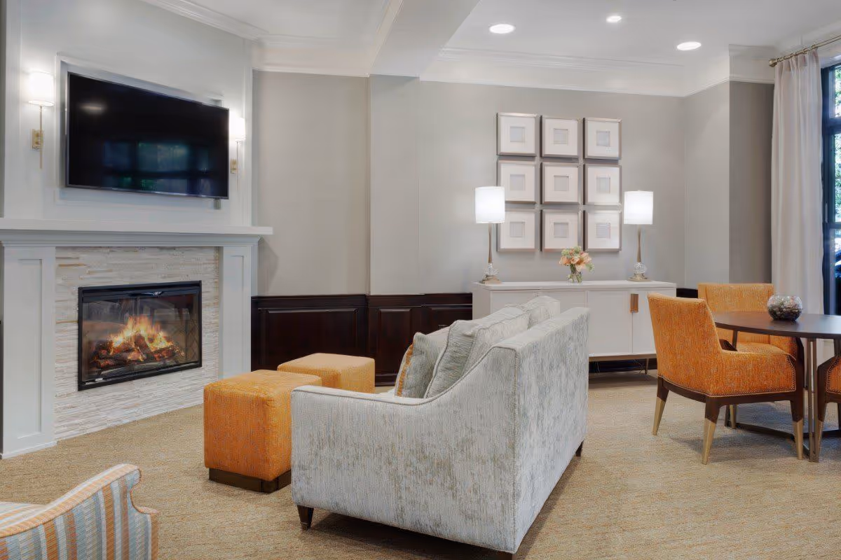 A cozy living room area with a light gray sofa facing a fireplace with a mounted flat-screen TV above it. Two orange ottomans are placed in front of the sofa. To the right, there is a round table with four orange upholstered chairs. On the far wall, a white sideboard with two lamps and a flower arrangement is visible beneath a grid of nine framed square artworks. Large windows with curtains allow natural light into the room.