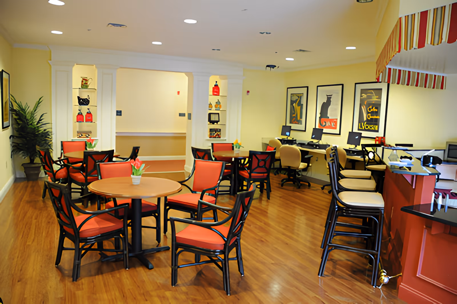 A bright, spacious room with several round tables surrounded by red cushioned chairs, a row of computer workstations with beige chairs against the wall, and a counter with high stools. The room has wooden flooring, yellow walls, decorative shelves with vases, framed artwork on the walls, and a potted plant in the corner.