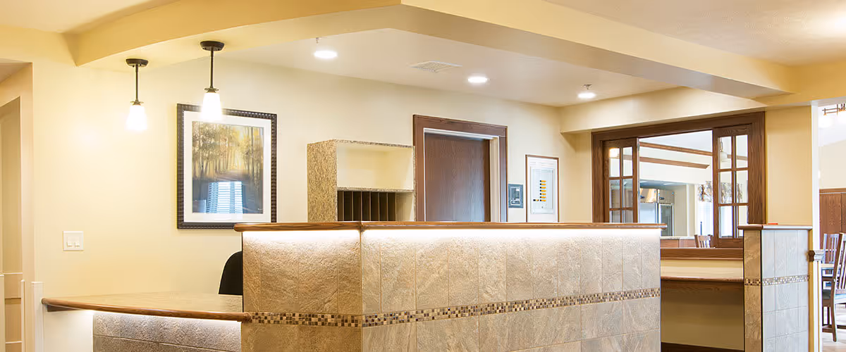 Reception area with a tiled front desk, two hanging pendant lights, a framed picture on the wall, and a doorway leading to another room with wooden framed windows and chairs visible.