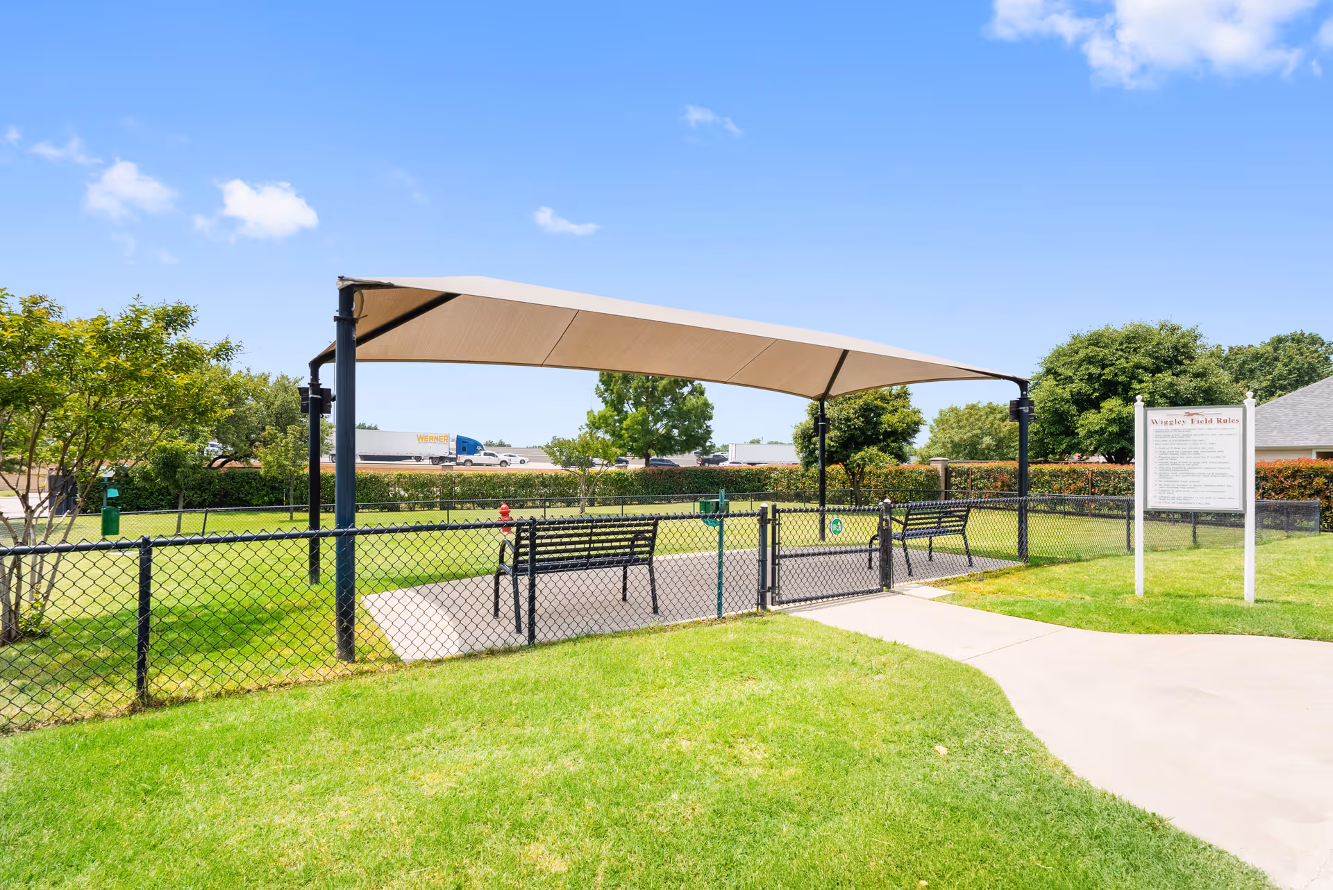 Outdoor fenced area with a shaded canopy covering benches on a concrete pad, surrounded by green grass and trees under a clear blue sky. A sign with rules is visible near the entrance of the fenced area.