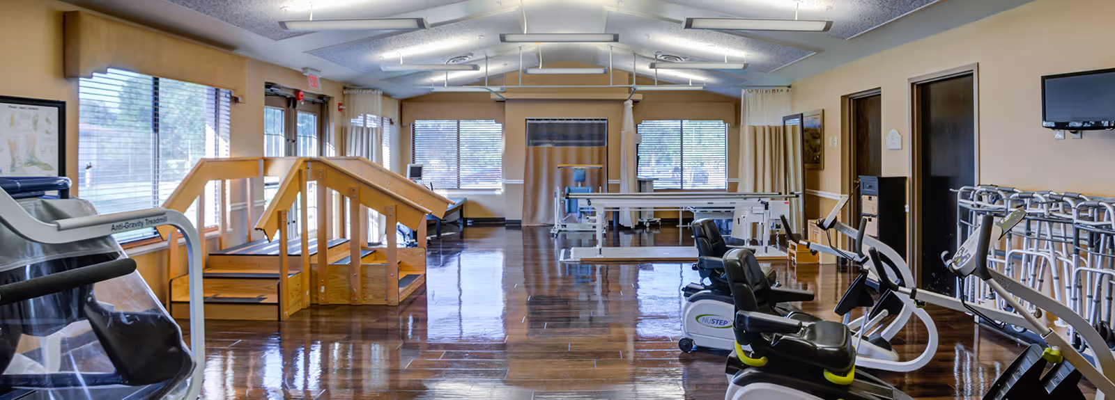 A spacious rehabilitation room with exercise equipment including stationary bikes, parallel bars, and wooden stairs for physical therapy. The room has large windows with blinds, polished wooden floors, beige walls, and ceiling lights.
