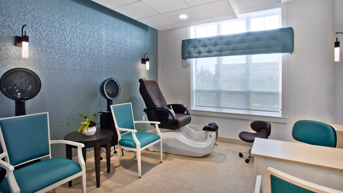 A modern salon room with two teal chairs, two hair dryers, a pedicure chair with a foot bath, a small table with a plant, and a desk with two office chairs. The room has a large window with a padded valance and patterned wallpaper on one wall.