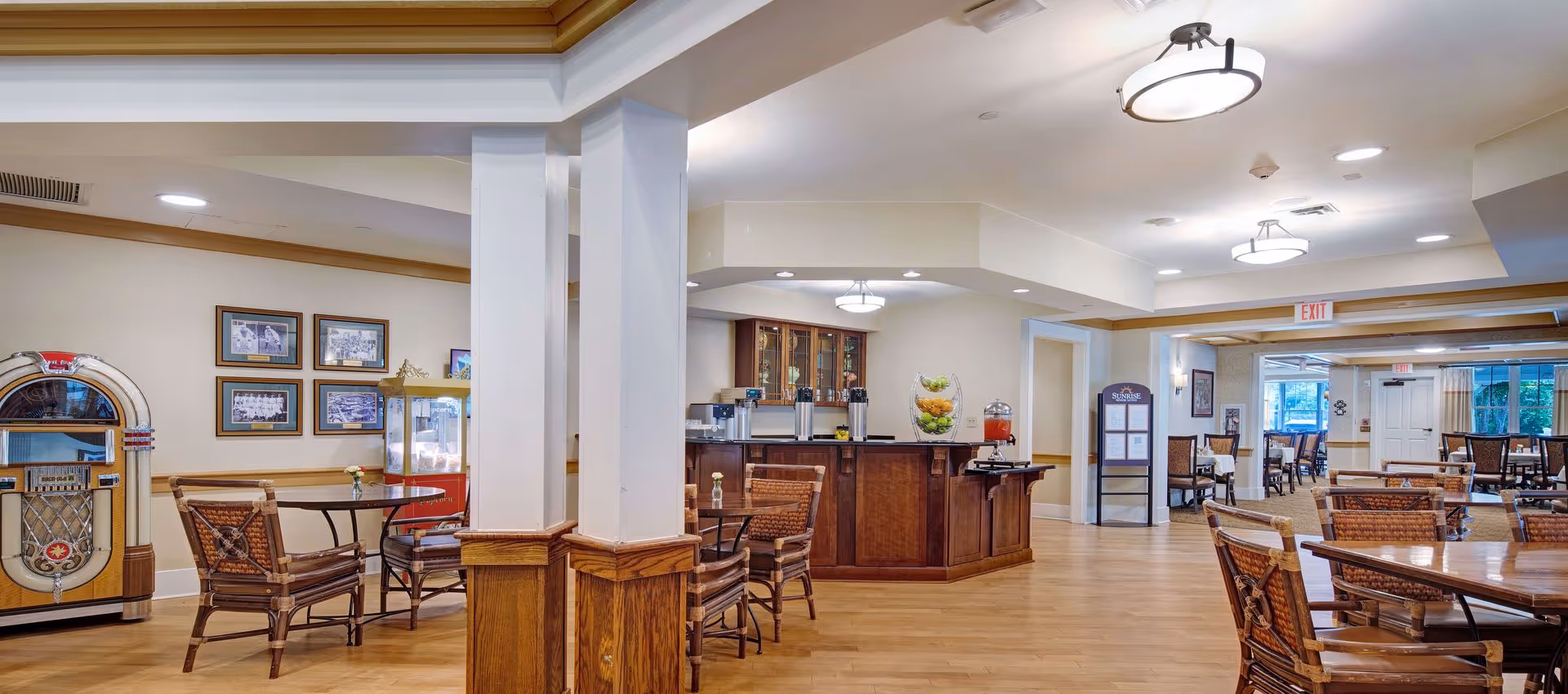 Spacious senior living dining area with wooden tables and chairs, a central beverage station/bar, and a jukebox.