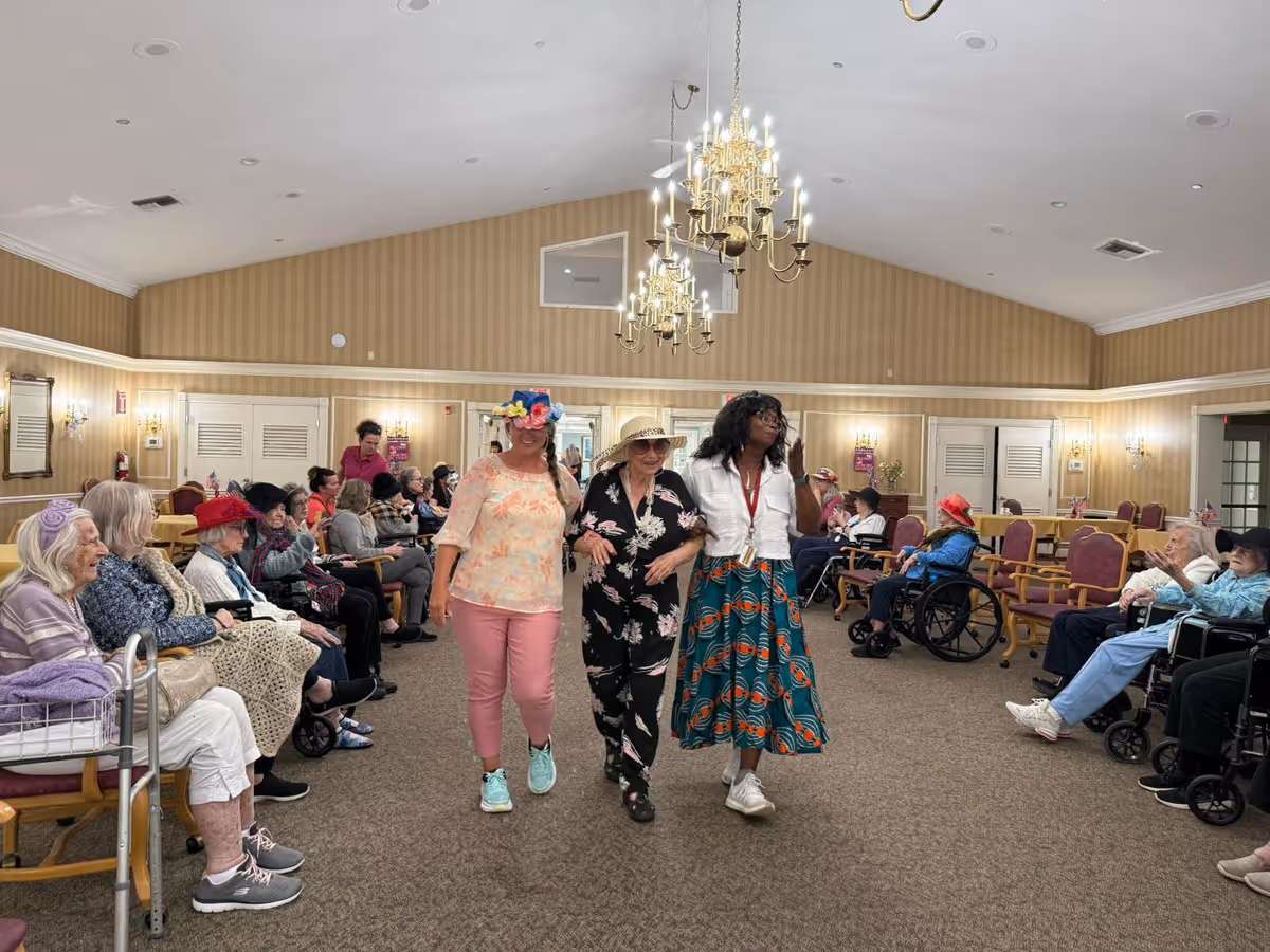 Three women walk down the center of a large senior living common room while seated residents watch from chairs and wheelchairs.