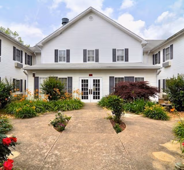 Front exterior view of a two-story white building with black shutters, surrounded by landscaped garden beds with various green plants and flowers, under a partly cloudy sky.