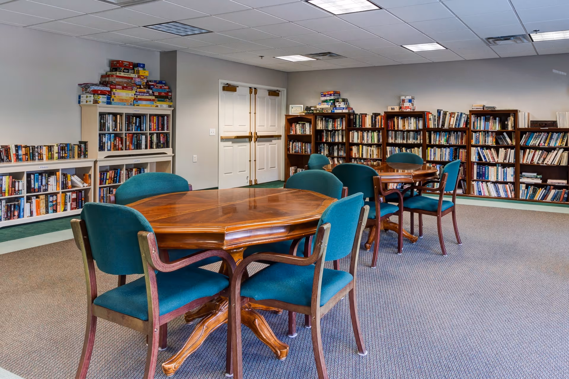 A quiet room with several wooden tables surrounded by teal upholstered chairs. The walls are lined with bookshelves filled with books and board games stacked on top. The room has a carpeted floor and a white double door in the background.