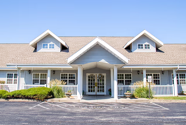 Front exterior view of a single-story assisted living facility building with a covered entrance supported by white columns, multiple windows, and a neatly maintained garden with shrubs and plants in front. The building has a light blue siding and a brown shingled roof with dormer windows.