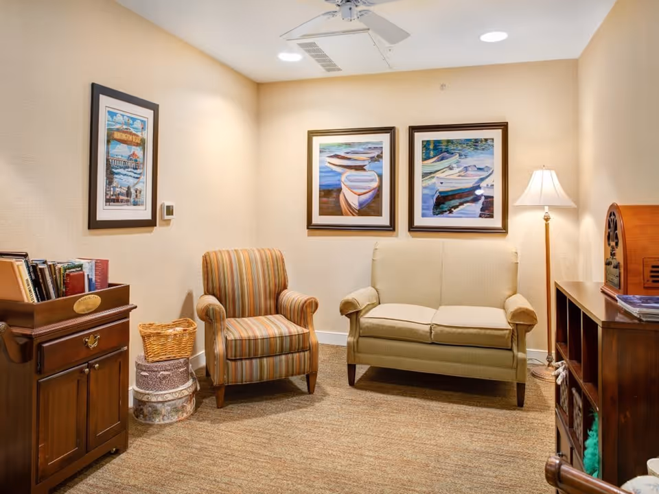 A cozy sitting area with a beige loveseat and a striped armchair. The walls are decorated with three framed paintings of boats and a Huntington Beach poster. There is a wooden cabinet with books and a basket on the left, and a floor lamp next to the loveseat on the right. The room has warm lighting and a carpeted floor.