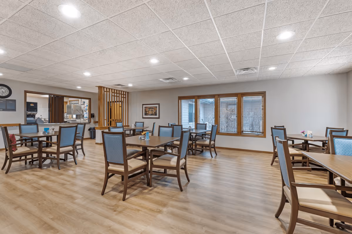 A spacious dining room in a senior living facility with multiple wooden tables and chairs arranged neatly. The room has light-colored wood flooring, a drop ceiling with recessed lighting, and large windows with wooden blinds letting in natural light. A serving window to the kitchen area is visible on the left side of the room.
