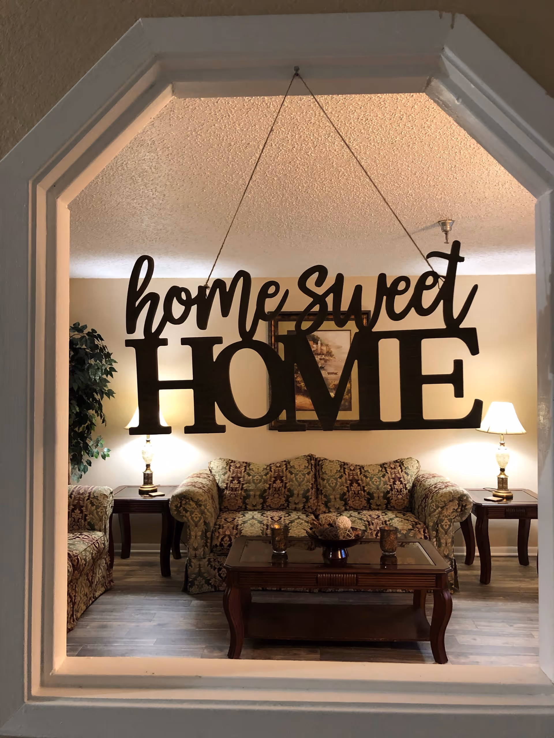 View through a window-like frame into a cozy living room with a patterned sofa and armchair, two side tables with lamps, a wooden coffee table with decorative items, a framed painting on the wall, and a hanging sign that reads 'home sweet HOME'.