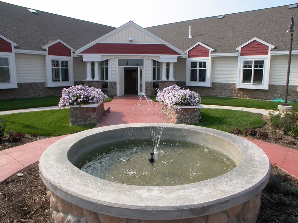 Outdoor view of the entrance to a building with a circular stone fountain in the foreground, water spraying from the fountain, flower beds with pink flowers on either side of the walkway leading to the open double doors, and a well-maintained lawn surrounding the area.
