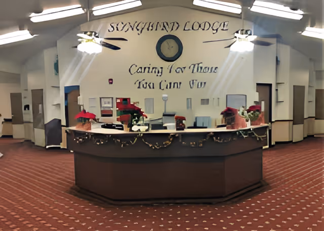 Reception area inside Songbird Lodge with a central desk decorated with poinsettia plants and garlands. The wall behind the desk displays a clock and the text 'SONGBIRD LODGE Caring For Those You Care For'. The room has a red carpet with a pattern and ceiling fans with lights.