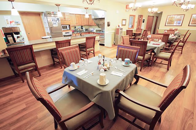 A dining room in a senior living facility with several tables covered with tablecloths and set with cups, plates, and utensils. The room has wooden flooring, upholstered chairs, and a kitchen area with appliances visible in the background.