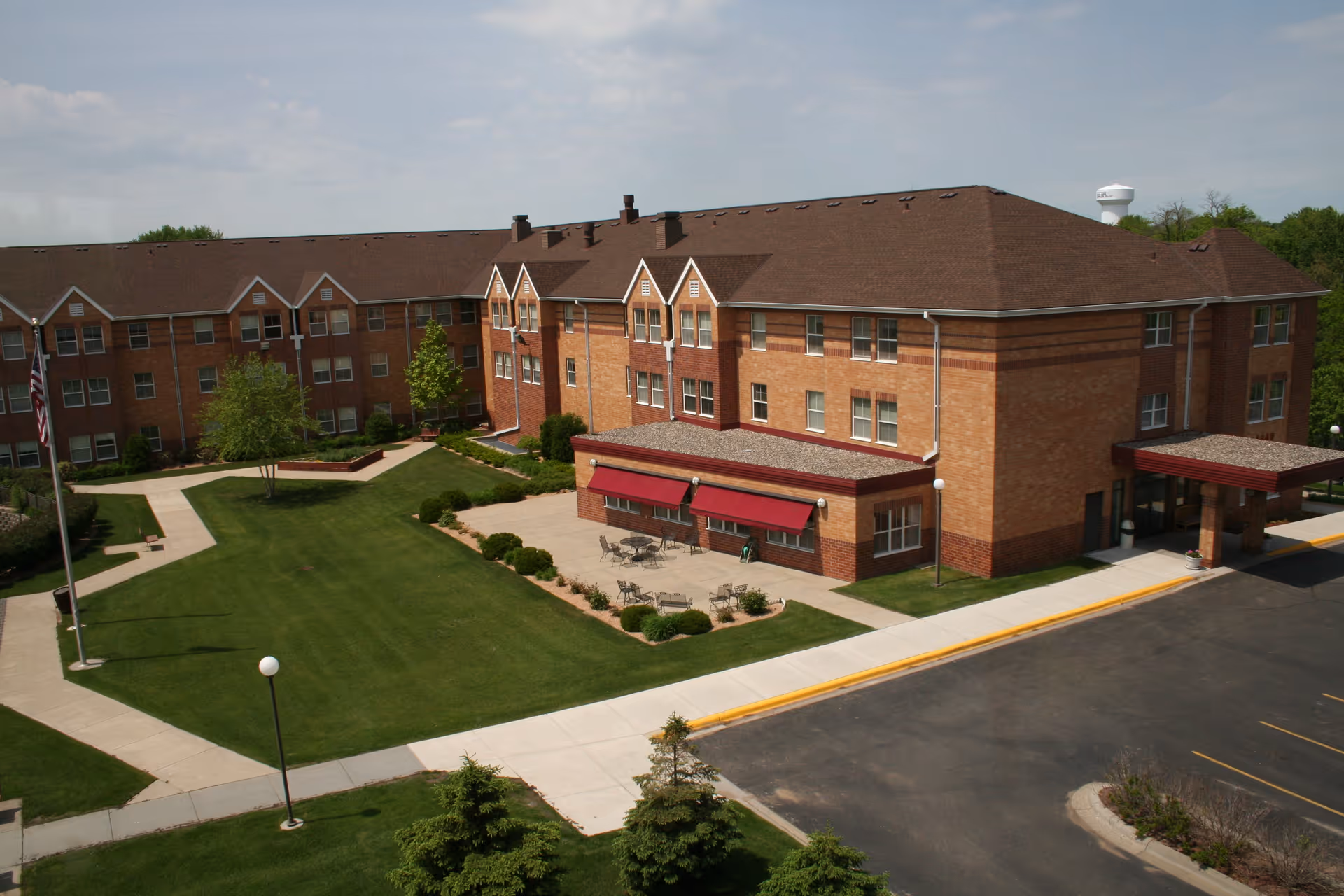 Brick three-story senior living facility building with a landscaped courtyard, patio seating, and adjacent parking area.