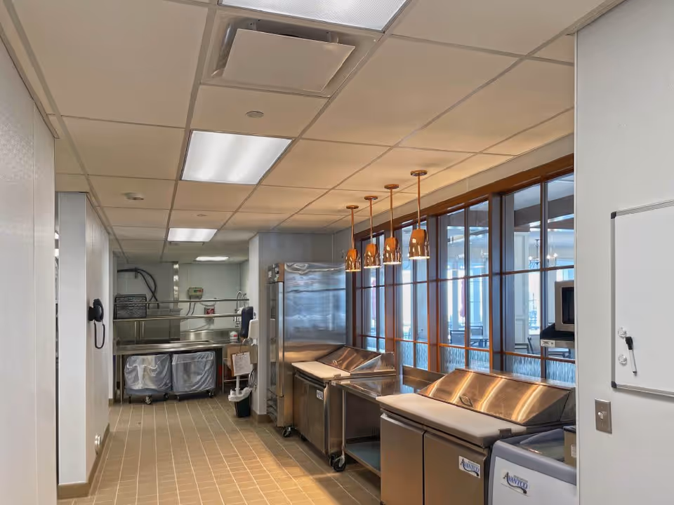 Commercial kitchen prep area with stainless steel counters, refrigeration units, pendant lights, and service windows.