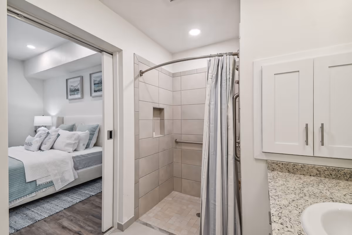 Tiled walk-in shower with curtain and grab bar in a bathroom opening to a furnished bedroom.