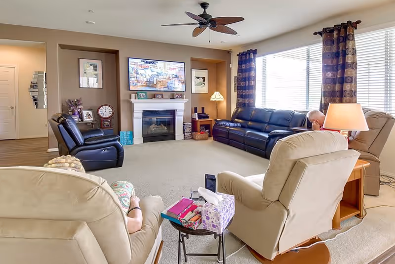 A cozy living room with beige carpet and neutral walls featuring a wall-mounted TV above a white fireplace. The room has a ceiling fan, large windows with patterned curtains, and various seating options including a black leather sofa, a black recliner, and beige recliners. Two elderly individuals are seated in the recliners, one partially visible. A small table with tissues and books is in the foreground.