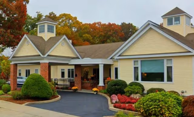 Exterior view of a single-story senior living facility building with beige siding, a covered entrance supported by white columns, well-maintained landscaping including bushes and flowers, and trees with autumn foliage in the background.