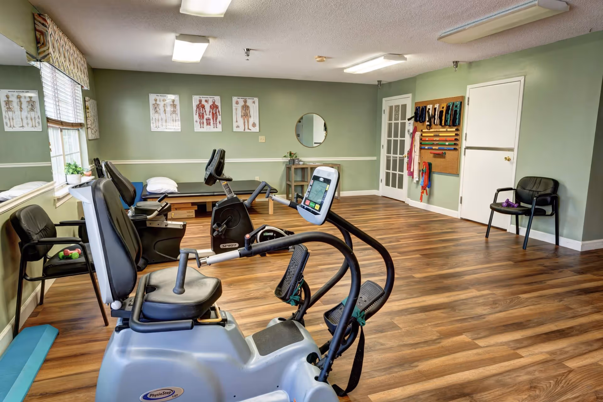 A fitness room with exercise equipment including a recumbent bike and a stationary bike. The room has wooden flooring, green walls, and anatomical charts on the wall. There are chairs along the walls and various exercise bands hanging on a pegboard.