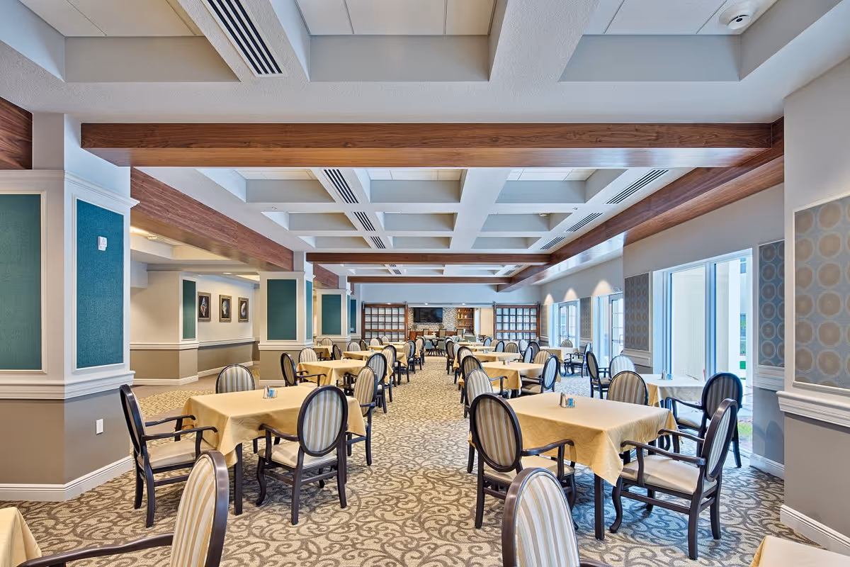 A spacious dining room with multiple tables covered in beige tablecloths, each surrounded by four upholstered chairs with striped cushions. The room features a patterned carpet, large windows on one side allowing natural light, and decorative wooden beams on the ceiling. The walls are adorned with framed artwork and decorative panels.