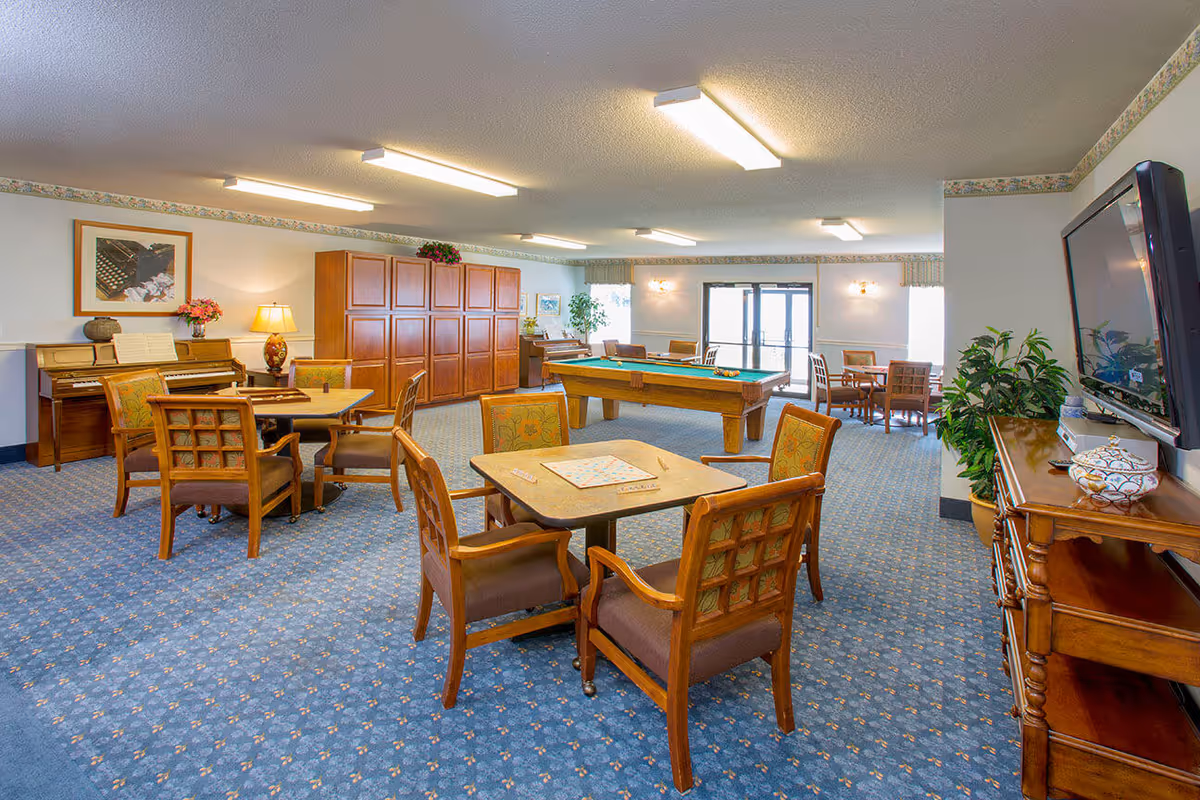 A spacious common room with blue patterned carpet featuring several wooden tables and chairs arranged for games and socializing. A pool table is centered in the room, with a piano and framed artwork on the left wall. Large windows and glass doors provide natural light, and a flat-screen TV is mounted on the right wall above a wooden console table with decorative items. Potted plants add greenery to the space.