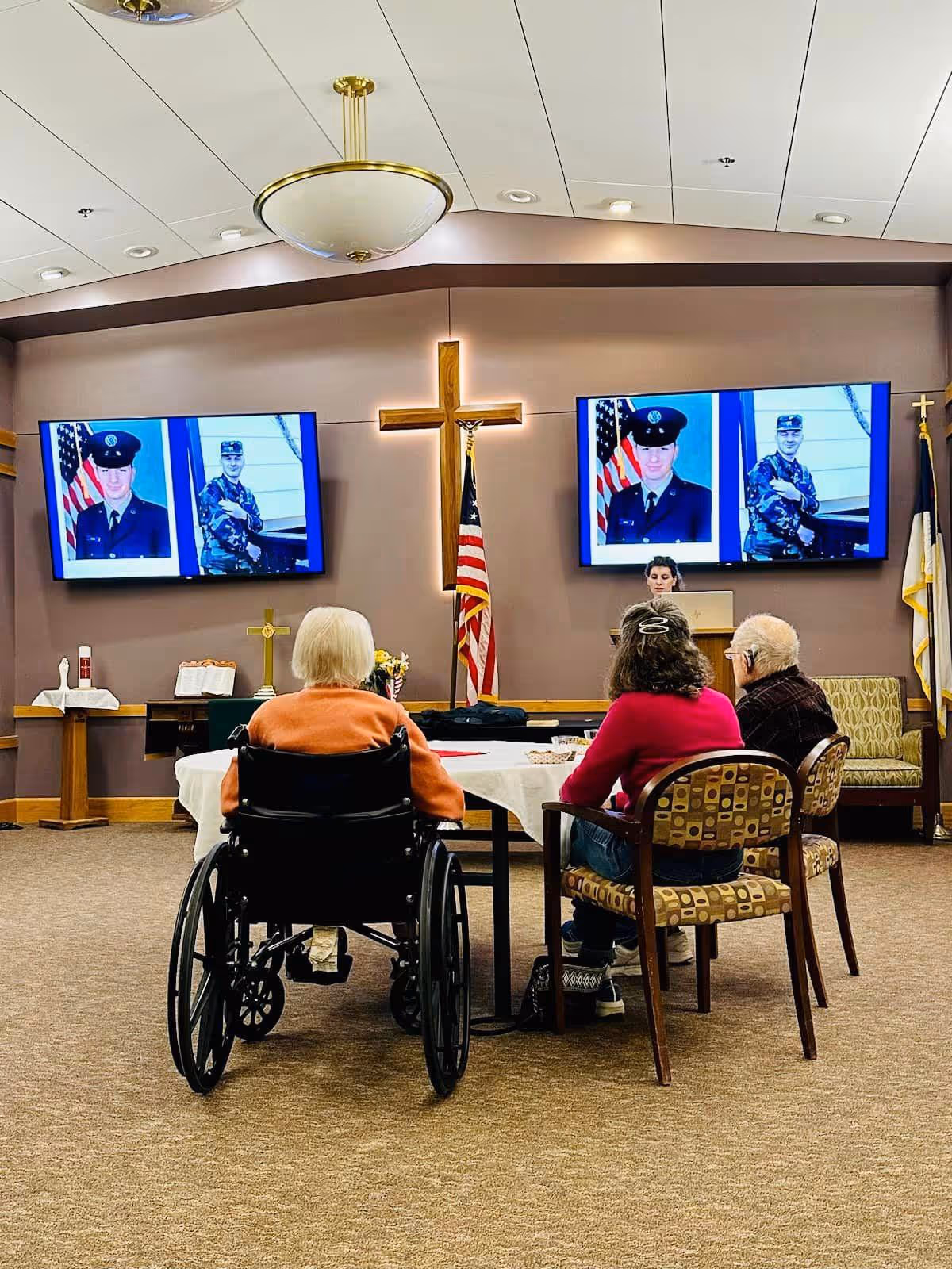 Three elderly people sitting at a table in a room with a large wooden cross and an American flag on the wall. Two large screens display images of a person in military uniform. A woman stands at a podium in front of the group.