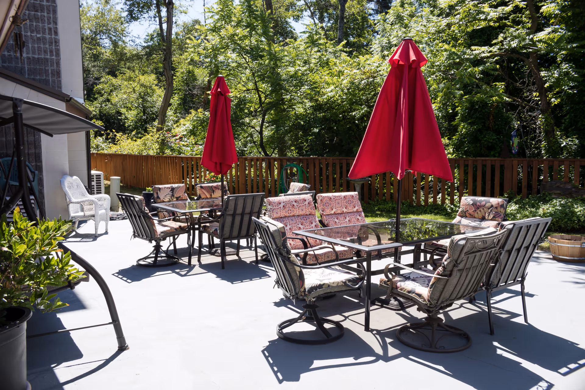 Outdoor patio area with two glass-top tables surrounded by cushioned chairs and two closed red umbrellas. The patio is adjacent to a building and bordered by a wooden fence with green trees and plants in the background.