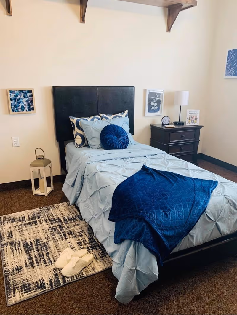 A neatly made single bed with light blue bedding and dark blue decorative pillows and throw blanket in a cozy bedroom. Next to the bed is a dark wooden nightstand with a lamp, clock, and framed picture. A patterned rug with white slippers is placed on the floor beside the bed. The walls are light-colored with framed artwork and a wooden shelf above the bed.