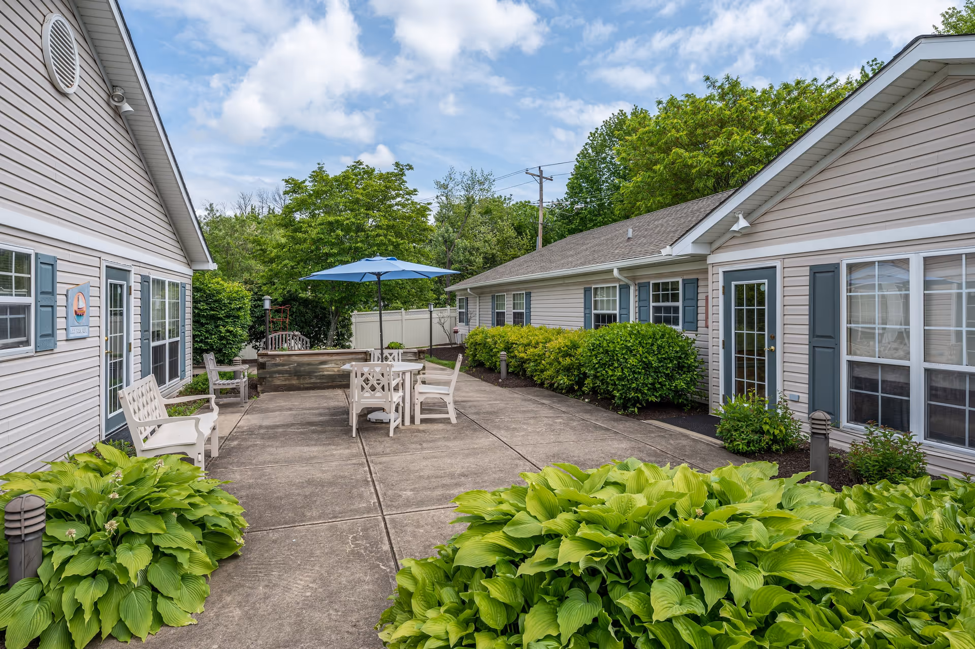 Outdoor patio area between two single-story buildings with beige siding and blue shutters. The patio has a concrete floor with white chairs and a table under a blue umbrella. Green bushes and plants surround the patio, and trees are visible in the background under a partly cloudy sky.