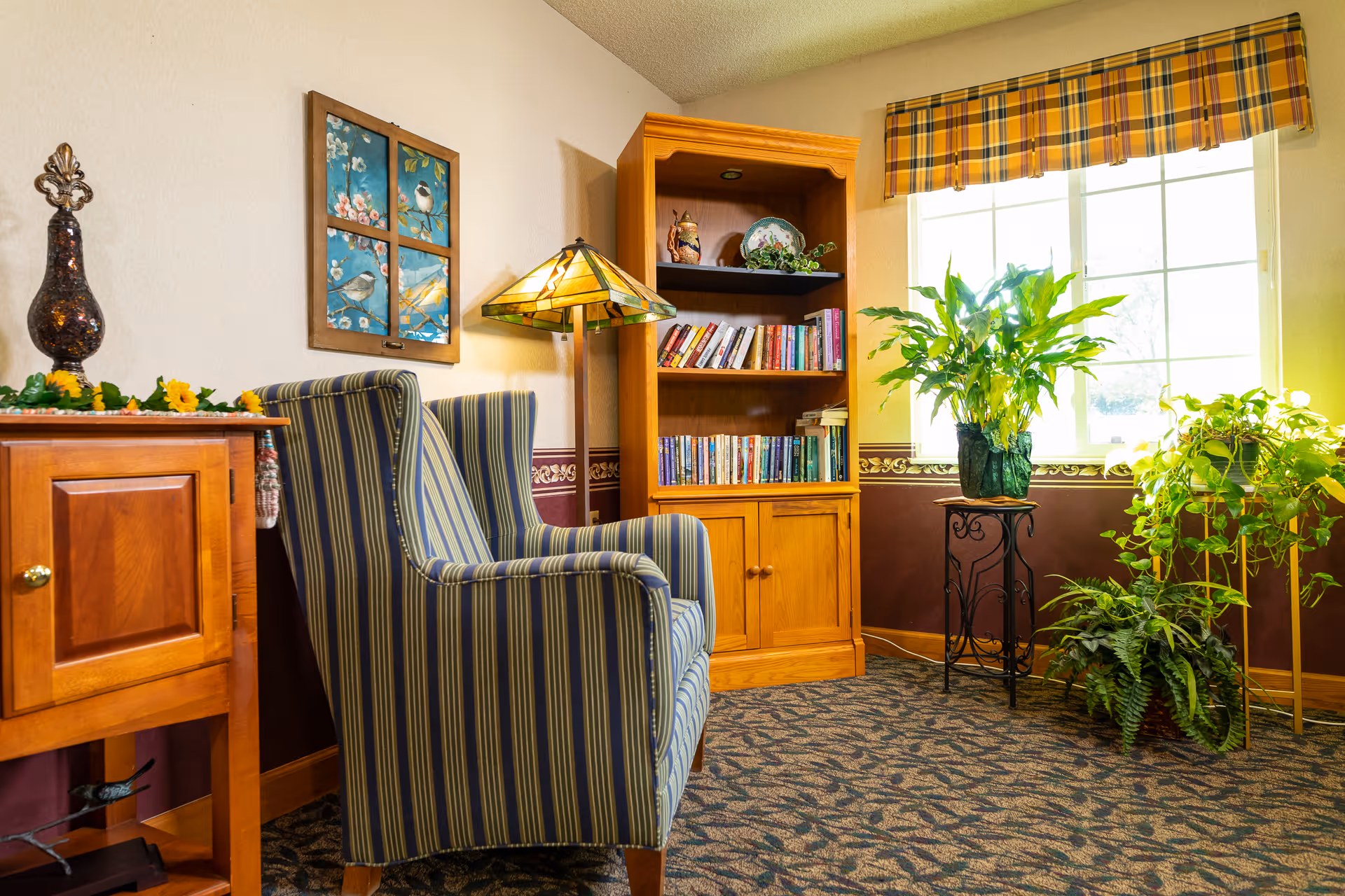 A cozy sitting area with a striped armchair, a wooden bookshelf filled with books, a Tiffany-style floor lamp, and several green plants near a window with a yellow plaid valance. The room has patterned carpet and a wooden cabinet with decorative items on top.