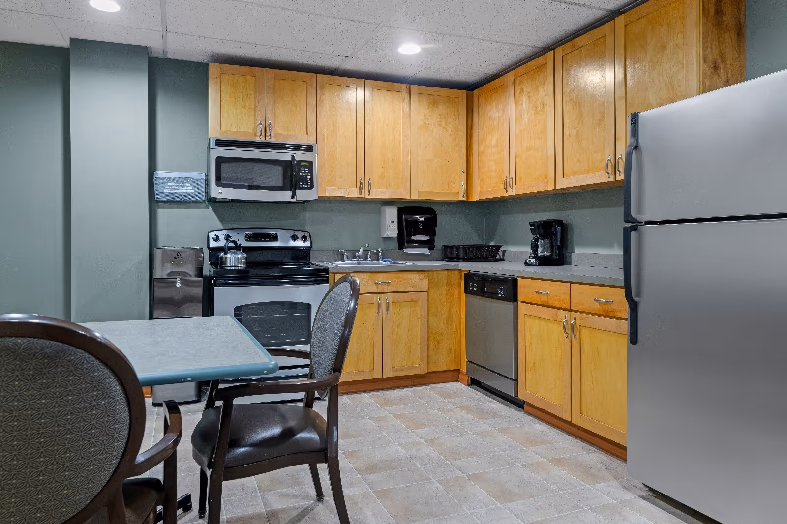 A kitchen area with wooden cabinets, a stainless steel refrigerator, microwave, stove, dishwasher, and coffee maker. There is a small table with two chairs in the foreground. The walls are painted green and the floor is tiled.