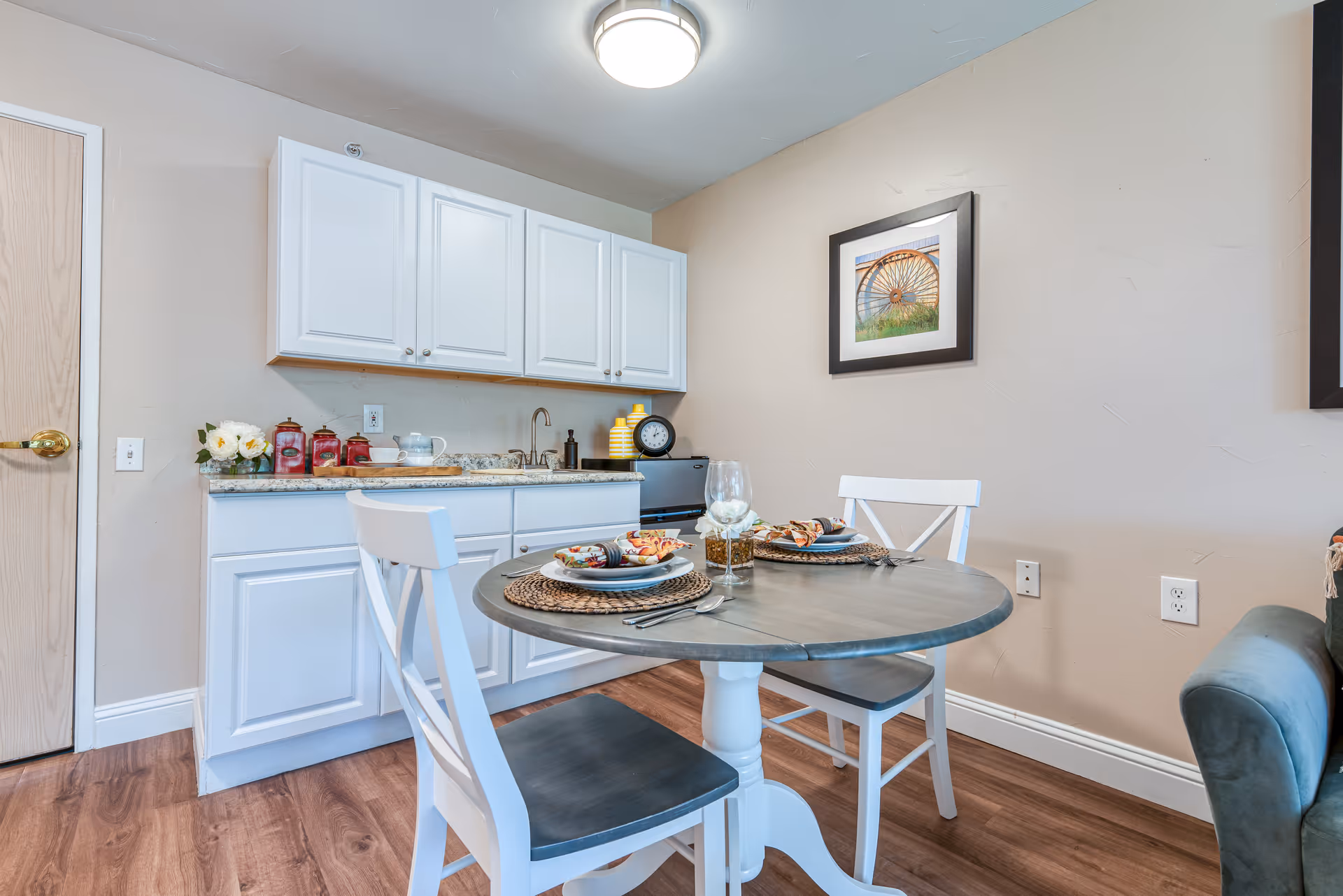 Small dining area with a round table set for two and white kitchenette cabinets in a senior living unit.